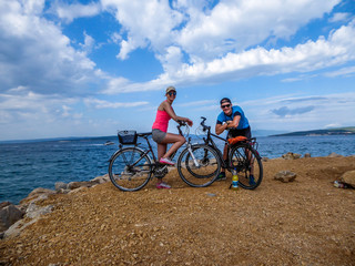 Fototapeta premium A couple in sporty outfits, standing on their bikes and checking the weather. Behind them there is Mediterranean Sea. Thick clouds above them. Active holidays. Beautiful coastal line of Croatia.