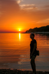 Fototapeta premium A girl enjoying the sunset by a beach. The sun sets over the horizon. The sun beams reflecting in the calm sea waters. There is an island on the side. The sky turns yellow. Girl enjoys the spectacle