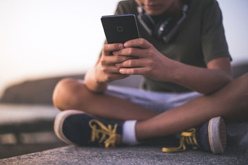 Close up view of a teen playing with smartphone. Young male sitting near the sea writing messages with cellphone. Trendy boy looking social video online. Focus on phone. Youth new technology concept.