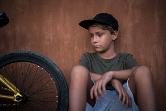Beautiful trendy boy relaxing alone sitting near his bicycle. Young man looking a bmx after riding on a summer day before start of the school. Portrait of a kid with rapper hat. Youth fun concept.