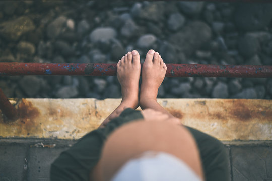 View from the top of a thoughtful teen sitting alone barefoot near the sea. Young boy with arms hold on to the knees think about future..Focus on feet. Youth, loneliness, adolescence problem concept