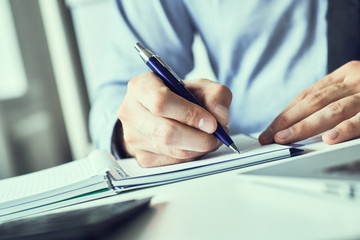 Businessman hand writing note on a notebook. Business man working at office desk. Close up of empty notebook on a blackboard with office supplies.