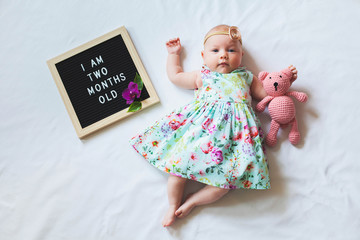 Two months old baby girl wearing floral dress and headband laying near text board and hugging teddy...