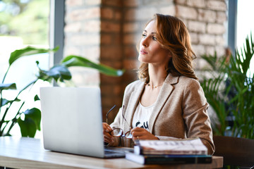 young woman in smart casual wear working on laptop while sitting near window in creative office or cafe