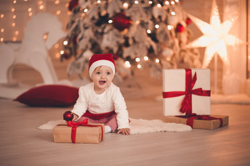 Christmas baby girl 1-2 year old wearing santa claus hat and suit open presents in room over tree with decorations. Winter holidays.