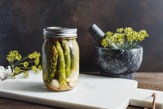 Pickled asparagus in a glass jar on a white marble tray. Seasonal canning vegetable recipe.