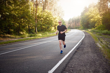 Caucasian young curly man athlete runs sunny summer day on asphalt road in the forest.