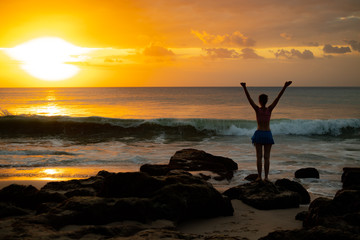 Fototapeta premium Excited young woman raising arms at the beach in front of the ocean. View from back. Sunset at the beach. Bali, Indonesia.