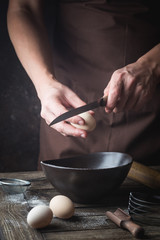 Professional chef hands are breaking an egg into bowl to make dough on wooden table, over dark background