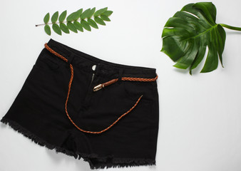 Black denim shorts with a leather belt on a white background with green tropical leaves. Top view