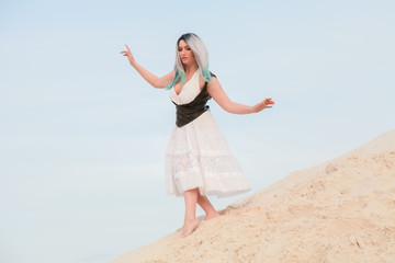 Obraz premium Young beautiful Caucasian woman in white dress and brown leather waistcoat posing in desert landscape with sand.