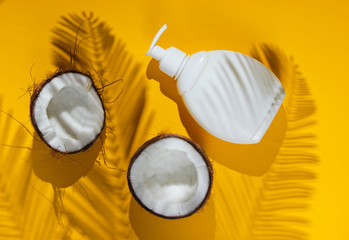 Minimalistic beauty still life. Two halves of chopped coconut and white bottle of cream with shadows from palm leaves on yellow background. Creative fashion concept. Top view.