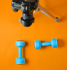 Fitness blogger. Sport concept. Plastic dumbbells and a camera on a tripod on orange background. Top view