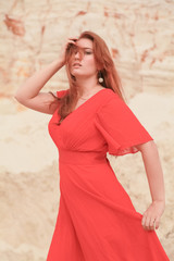 Young beautiful Caucasian woman in long red dress posing in desert landscape with sand.