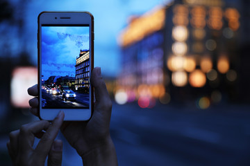 Woman taking photo of modern city in evening with smartphone, focus on hand. Bokeh effect - obrazy, fototapety, plakaty