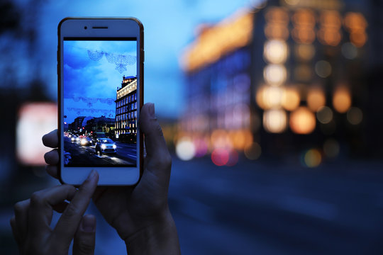 Woman taking photo of modern city in evening with smartphone, focus on hand. Bokeh effect