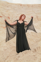 Young beautiful Caucasian woman in black chiffon dress posing in desert landscape with sand.