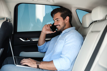 Attractive young man working with laptop and talking on phone in luxury car