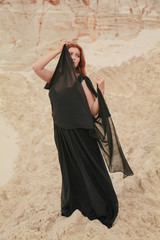 Young beautiful Caucasian woman in black chiffon dress posing in desert landscape with sand.