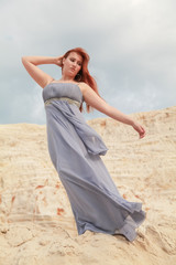 Fototapeta na wymiar a red-haired girl in a long grey dress fluttering in the wind stands on a mound of sand in the desert and loves the wind
