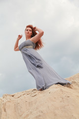 a red-haired girl in a long grey dress fluttering in the wind stands on a mound of sand in the...