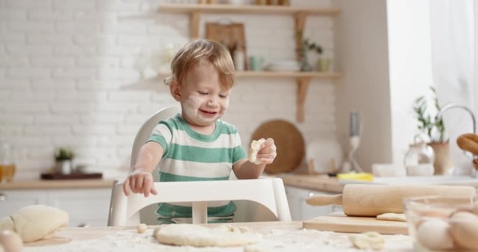 Cute little baby left alone in kitchen, messing with dough with face covered with flour. Child playing with food and positively smiling close up
