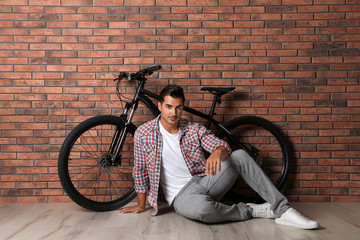 Handsome young man with modern bicycle near brick wall indoors