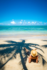 Fototapeta na wymiar Relaxed tourist wearing a straw sun hat reclining on the shore of a sunny palm-lined tropical island beach 