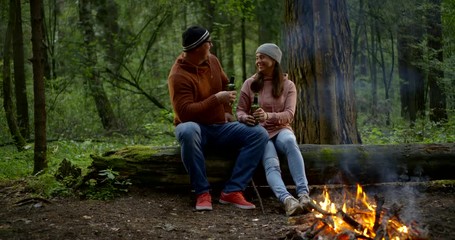 man and woman are sitting on log near fire in forest, holding beer bottles and chatting cheerfully