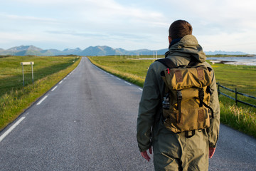 Fototapeta premium Man with backpack walking on an empty road. Amazing landscape, mountain and ocean. Scenic view. New way. Enjoy the moment, relaxation. Wanderlust. Travel, adventure, lifestyle. Explore North Norway