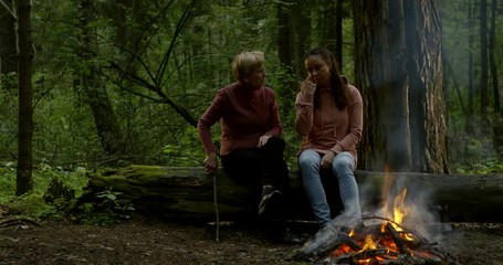 elderly mother and adult daughter are chatting cheerfully resting together in forest near campfire