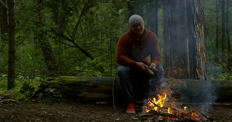 lonely adult hiker is making fire in forest in camp during halt, throwing firewoods