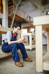 Full length portrait of young female worker using smartphone while resting on wooden stairs during break, copy space