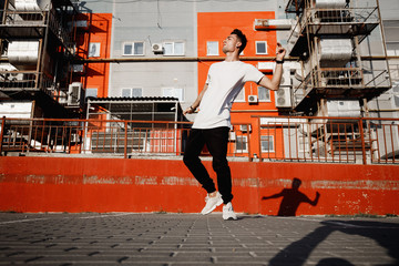 Young guy dressed in jeans and t-shirt is dancing modern dance in the street on the background of urban buildings in the warm day