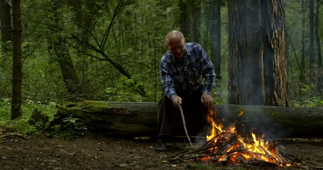 mature hiker is resting in forest sitting on log near burning fireplace in daytime