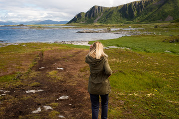 Naklejka premium Blond hair girl walking, looks forward and enjoys a stunning scenic view. Ocean and rocks, beautiful landscape. Wanderlust. Adventure, freedom, lifestyle. Explore North Norway. Summer in Scandinavia