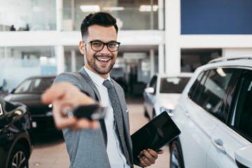 Good looking, smiling salesman poses in a car salon or showroom. He stands with outstretched hand...