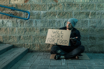 A man, homeless, a man asks for alms on the street with a sign will work for food. Concept of homeless person, addict, poverty, despair.