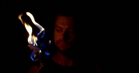 A man stands in the dark with two torches of fire in his hands and illuminates his face meditating on the flames