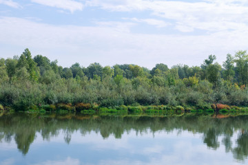 Beautiful summer forest riverside view landscape. Sandy beach on the shores, green grass and trees, blue river and sunny cloudy sky. Concept for leisure, travel, tourism
