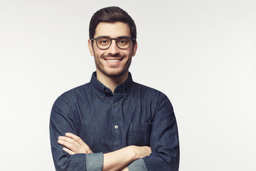 Young man with crossed arms wearing glasses, looking at camera with smile, standing isolated on gray