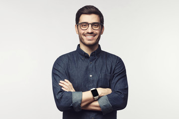 Man with crossed arms wearing glasses and smart watch, looking at camera with smile, standing...