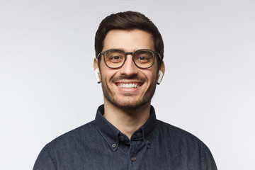 Close-up headshot of young smiling man wearing blue denim shirt, listeting to his favorite music track with earphones