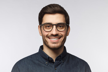 Close-up portrait of young man feeling happy and smiling, wearing denim shirt and stylish glasses, isolated on gray background