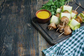 Skewers on a skewer with zucchini with sauce, vegetables and herbs on a cutting board. Dark wooden background.