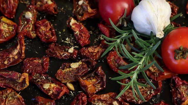 Sun-dried tomatoes with spices and garlic on a baking sheet. Nearby are fresh rosemary, tomatoes and a head of garlic.