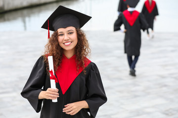 Happy African-American student in bachelor robe and with diploma outdoors