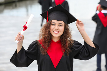 Happy African-American student in bachelor robe and with diploma outdoors