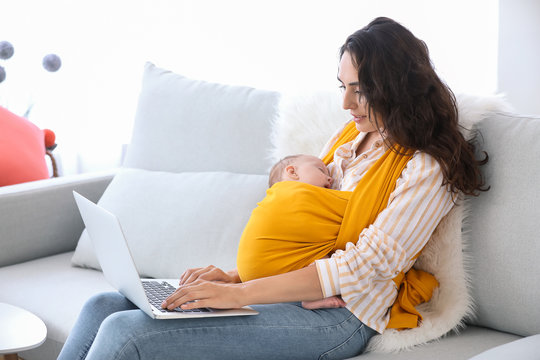 Young mother with little baby in sling using laptop at home - Powered by Adobe