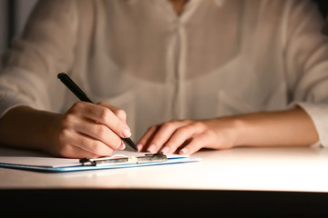 Woman writing something on sheet of paper at workplace in evening, closeup